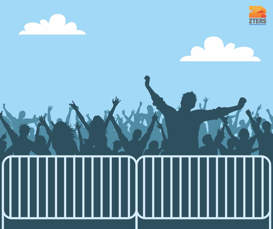 A large group of people with their arms up at a concert. A crowd control barrier is in front of them. There are clouds in the sky. The ZTERS logo is in the top right.