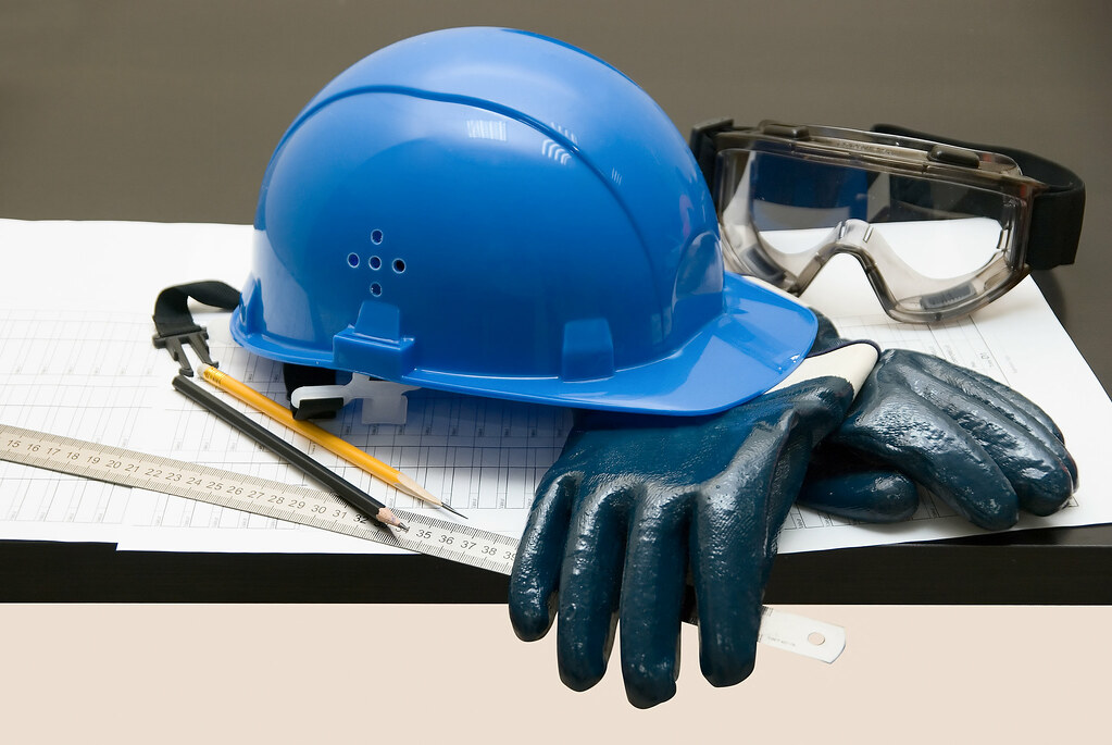 A picture of a pile of safety gear (gloves, goggles, and a hard hat) on top of a pile of documents, measuring stick, and pencils.