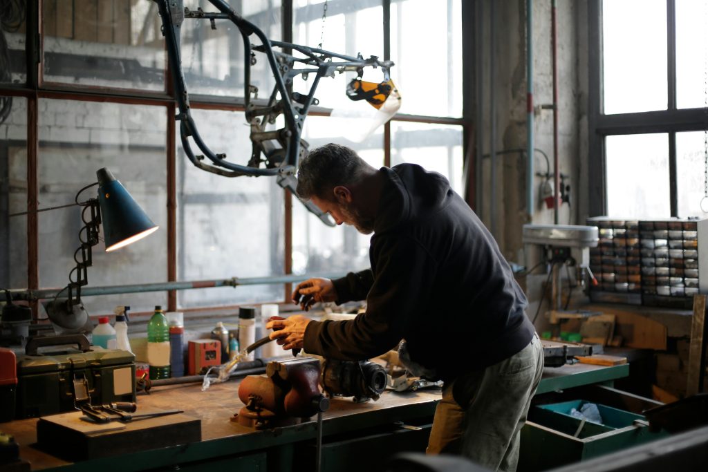 A picture of a man in a workshop using tools connected to a extension cord with various work lighting equipment above him.